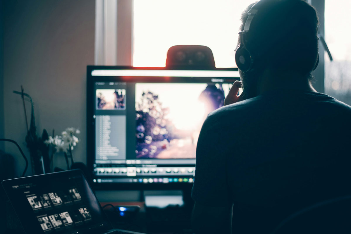 Un uomo che indossa cuffie e guarda un monitor mentre modifica foto in un ambiente luminoso. Sul desktop ci sono immagini aperte.