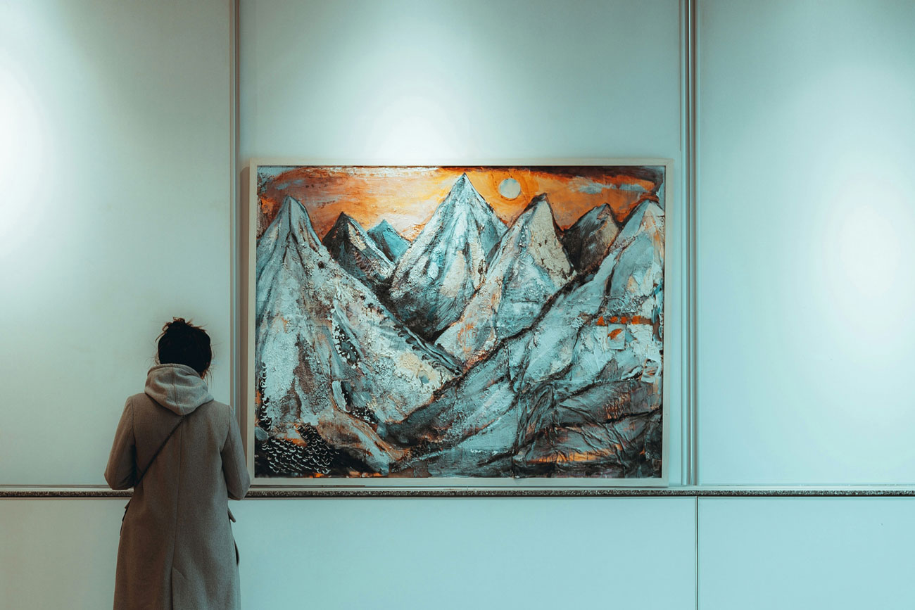 Una persona osserva un dipinto di montagne in una galleria d'arte, illuminata da luci soffuse. Le vette si stagliano contro un cielo arancione.
